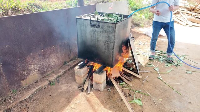 Outdoor scene of boiling Banh Chung in a large metal pot over a firewood flame. Showing the step of filling water, maintaining the fire for boiling Chung cake during Vietnamese Tet cooking tradition