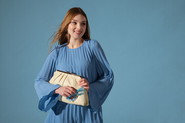 Beautiful fashionable happy smiling woman wearing trendy blue color pleated short dress, holding stylish leather pouch bag. Waist up studio portrait. Copy, empty space for text © Victoria Fox