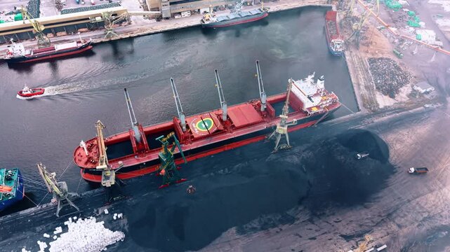 Cargo ship being loaded with coal at a port terminal. Heavy cranes operate over a large vessel docked next to piles of black minerals and an approaching tugboat.