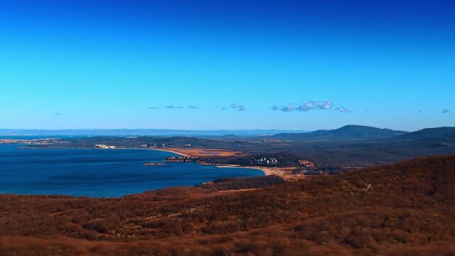 Panoramic view of a coastal sea bay with hills. Forested peninsulas extend into the calm blue water under a clear sky during daytime.