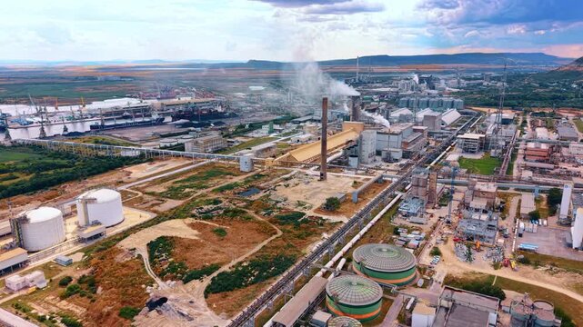 Industrial chemical refinery plant near a river terminal. White storage tanks and production units emit smoke within a large infrastructure zone next to distant water.