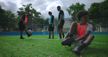 Fototapeta premium Young African American man tying shoelaces before casual soccer match with friends, preparing for game on sunny outdoor field, teamwork, focus, anticipation, and sports routine