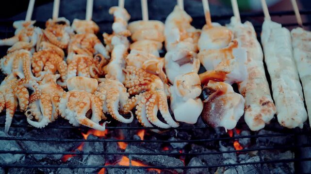 Close up of vendor grilling fresh squid skewers on hot charcoal stove in night market in Thailand.
