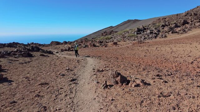 A solitary hiker walks along a rugged volcanic trail from Pico Teide to Pico Viejo Tenerife, Canary Islands. The expansive, bleak landscape with distant ocean offers a sense of remote adventure.