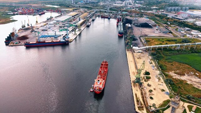 Red tanker ship navigating through a busy industrial port channel. An aerial photograph captures an oil vessel moving between concrete docks equipped with heavy cargo cranes.