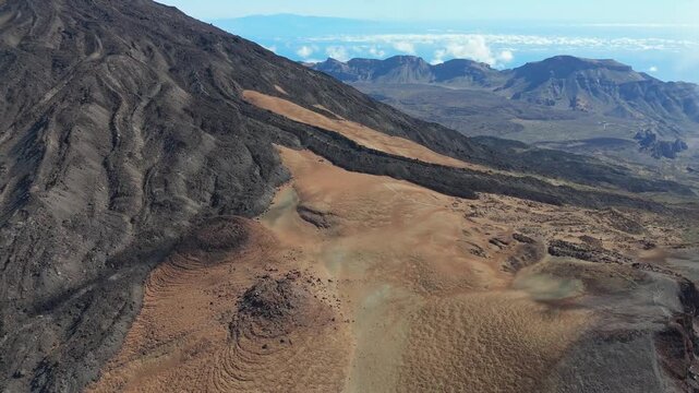 Aerial drone shot of the volcanic landscape of Pico Viejo on Tenerife. The drone pans left and booms up, revealing rugged terrain, sandy slopes, and distant ocean views.
