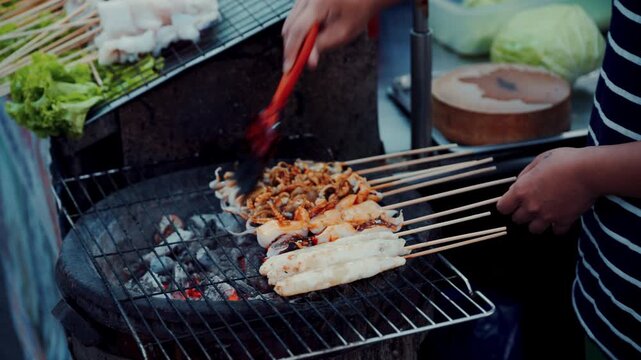 Close up of vendor grilling fresh squid skewers on hot charcoal stove in night market in Thailand.