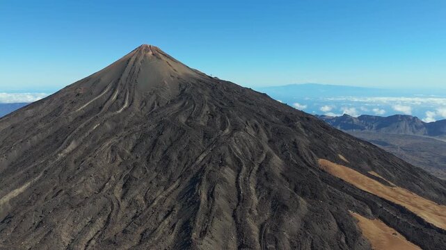 An aerial drone shot pans left and booms up over the majestic Teide volcano on Tenerife, showcasing its rugged, rocky terrain and the vast, remote volcanic landscape under a clear blue sky.