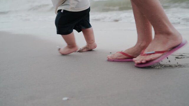 Close up of baby bare feet walking on wet sand beach with mother.