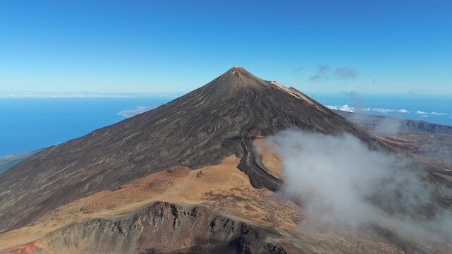 Aerial drone pans left, showing volcano Pico del Teide on Tenerife, Canary Islands. Dramatic volcanic landscape with rugged peaks, rocky terrain, and distant ocean views under a clear blue sky.