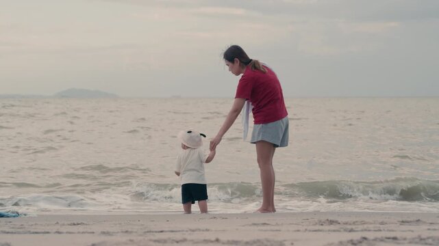 Asian mother holding hands with her baby standing on beach near ocean waves.