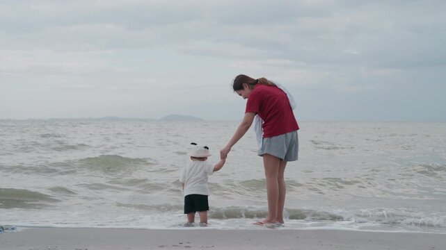 Asian mother holding hands with her baby standing on beach near ocean waves.