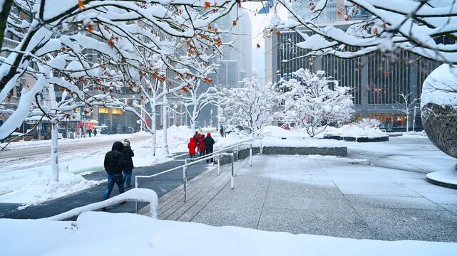 Snow covered trees and skyscrapers in New York City. People walk on a snow covered plaza surrounded by snow laden trees and high rise buildings during winter.