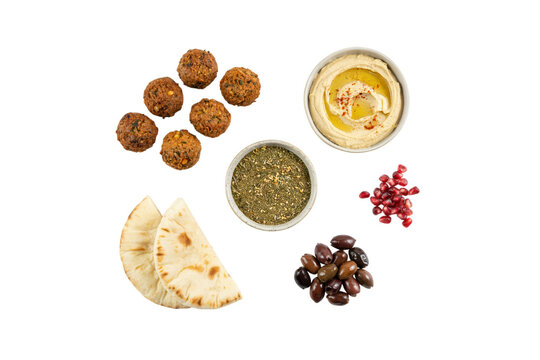 Middle Eastern mezze selection featuring falafel balls, hummus bowl, pita bread slices, black olives, pomegranate seeds, and zaatar spice on white