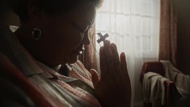 Side view closeup of senior Black woman praying with closed eyes while holding small wooden cross at home. Footage is ideal for religious media, spiritual counseling, and mental health campaigns
