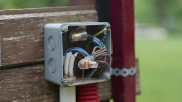 Electrical junction box with exposed wires and fuses mounted on wooden fence, showcasing detailed connections and components in a green outdoor setting
