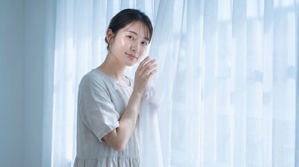 Fototapeta premium Young woman touching white curtains. Female standing by window in soft morning light.