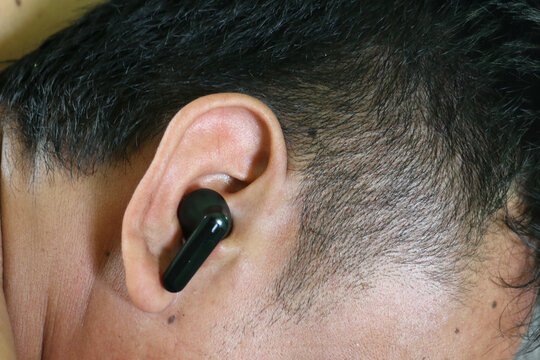 Close-up of a man wearing a black wireless earbud while relaxing. Ideal for concepts related to wireless audio, Bluetooth technology, hands-free communication, music streaming, podcasts, remote work, 