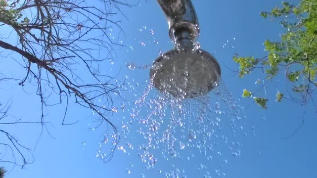 Horizontal HD 1080p super slow motion low angle shot of an outdoor metal shower head turning on and spraying fresh water against a clear blue sky in summer.