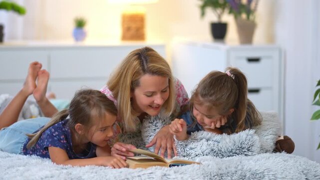 Loving mother reads a book to her two young daughters lying on the bed before bedtime