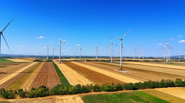 Wind farm stands on striped agricultural fields under blue sky. Drone shot captures white turbines installed along farmland rows under sunny atmosphere.