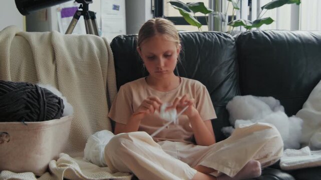 Focused young girl sits on a sofa in a bright home living room while crocheting soft white yarn with a small metal hook tool.