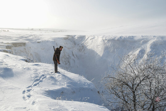 A hunter with a shotgun stands on the very edge of a deep snow-covered ravine on a winter day.