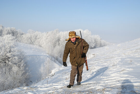A hunter in warm winter clothes and a fur hat walks along a snow-covered slope, holding a shotgun.
