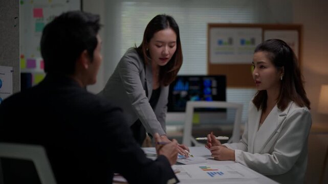 Asian colleagues brainstorming and collaborating during a late-night office meeting, analyzing financial reports and marketing data for a new business strategy