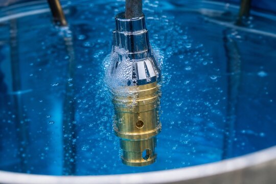 A brass component submerged in a blue electroplating bath, bubbles rising.