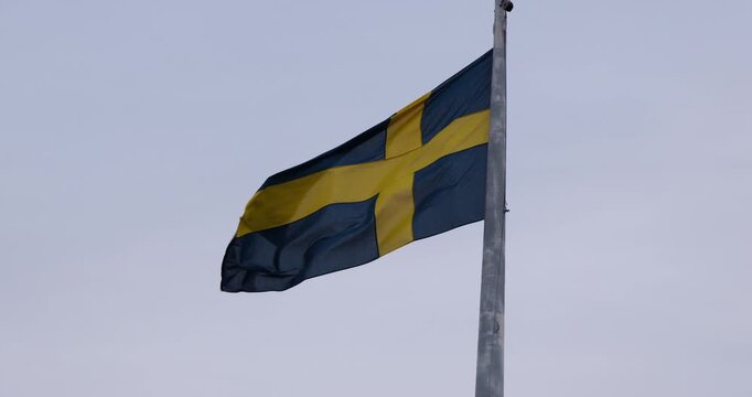 Low angle view of the Swedish national flag waving on a flagpole against an overcast sky.	
