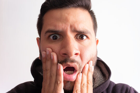 Portrait of a young Latino man, impressed and with his mouth open, staring at the camera against a white background in a photography studio. Concept of facial expression and strong emotions