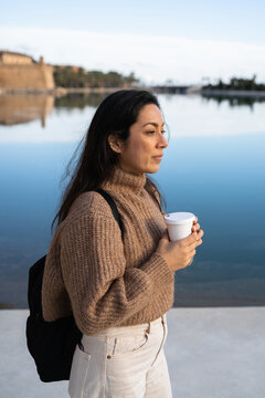 A woman standing by the water with a cup of coffee to go in her hand
