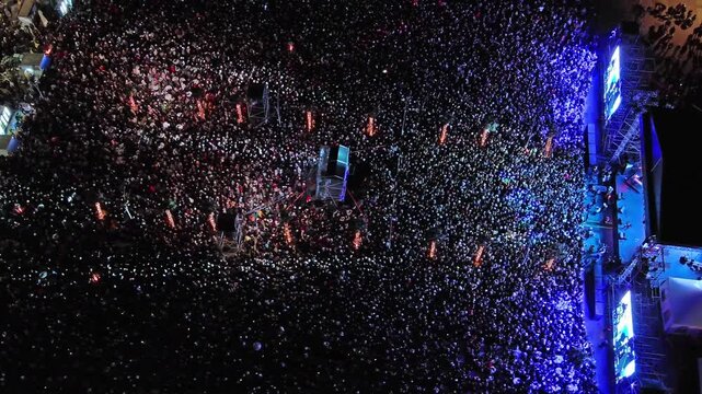Aerial around view of crowd at night music concert. People gathered at festival outdoor stage with lights and screens, 4k
