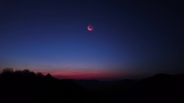 Blue hour time with lunar eclipse, stars and planets above landscape silhouettes.