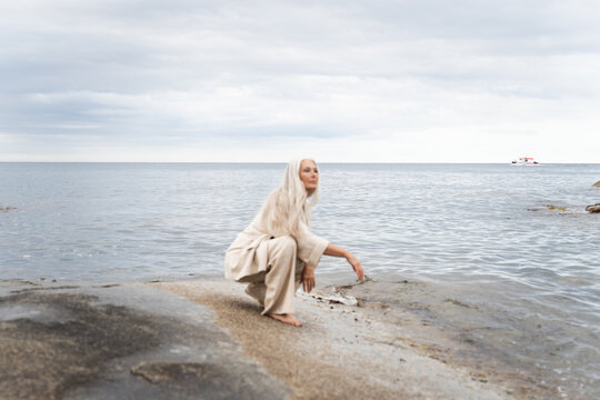 Woman Crouches by the Water on a Rocky Shore With Clouds Above