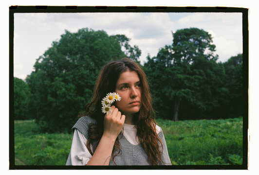 A young woman holds daisies beside her face in a green field