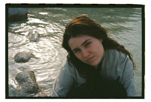 A young woman leans toward the camera by a rocky stream