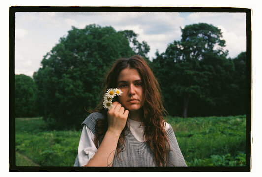 A young woman holds daisies beside her face in a green field