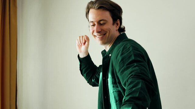 Joyful young man in a green shirt dancing energetically indoors. He smiles and laughs while making carefree dance moves against a white wall. High quality 4k footage