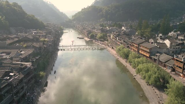 Horizontal 4K UHD aerial drone shot flying forward and tilting down over Tuojiang River stepping stones footbridge surrounded by traditional wooden houses in Fenghuang Ancient Town Hunan China