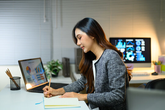 Business Woman Taking Notes While Reviewing Data Presentation on Laptop and Monitor in Modern Office