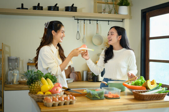 Two Happy Young Women Cooking Together in a Modern Home Kitchen with Fresh Vegetables and Fruits