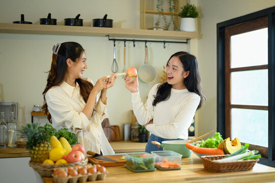 Two Happy Young Women Cooking Together in a Modern Home Kitchen with Fresh Vegetables and Fruits