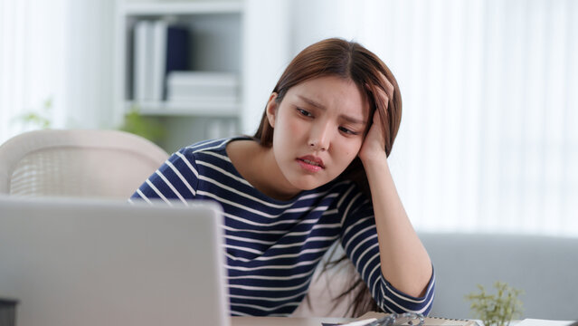 Stressed young Asian woman holding head in frustration while working on laptop at home with copy space
