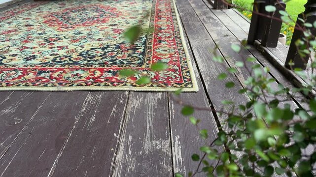 Close-up of a woman unrolling a vintage oriental rug across a weathered wooden porch floor during a cozy veranda makeover. Traditional patterned carpet adds warmth and character to an outdoor living 