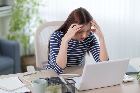 Stressed Asian woman holding head in frustration while working on laptop at home desk with copy space
