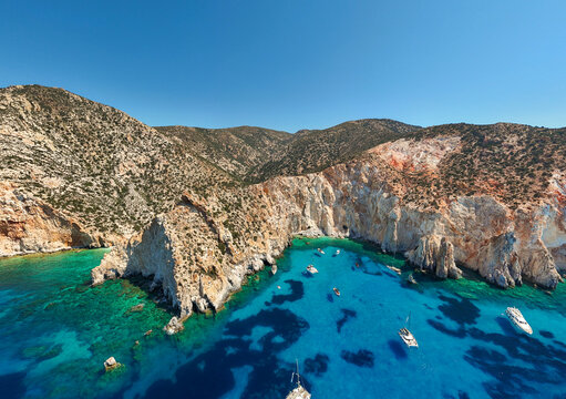 Aerial view of Kleftiko Bay on Milos Island, Greece, showcasing dramatic white cliffs, turquoise water, and anchored boats beneath a clear summer sky &mdash; a stunning example of Aegean coastal beauty.