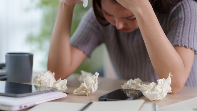 Stressed Asian woman holding tissue with crumpled papers on desk expressing frustration at home workspace