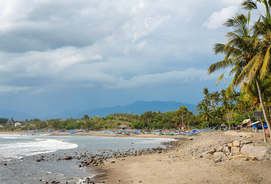 Scenic view of Medewi Beach in Bali, Indonesia, featuring traditional fishing boats, a tropical coastline, swaying coconut palms, distant mountains, and dramatic clouds.